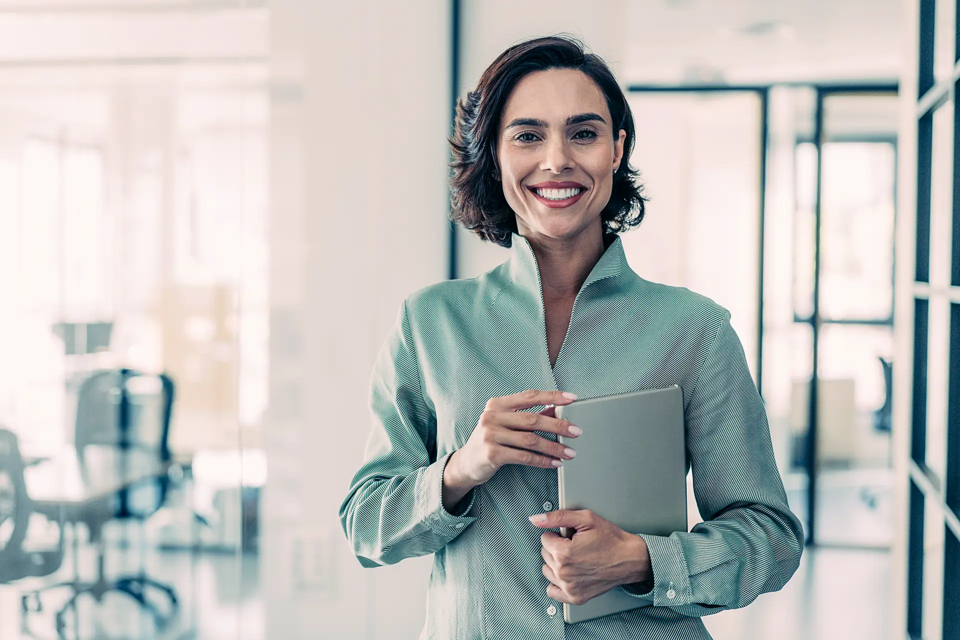 Eine lächelnde Person in einem hellgrünen Oberteil steht in einem modernen Büro und hält ein Tablet in der Hand. Im Hintergrund sind Glaswände und ein Konferenzraum zu sehen.