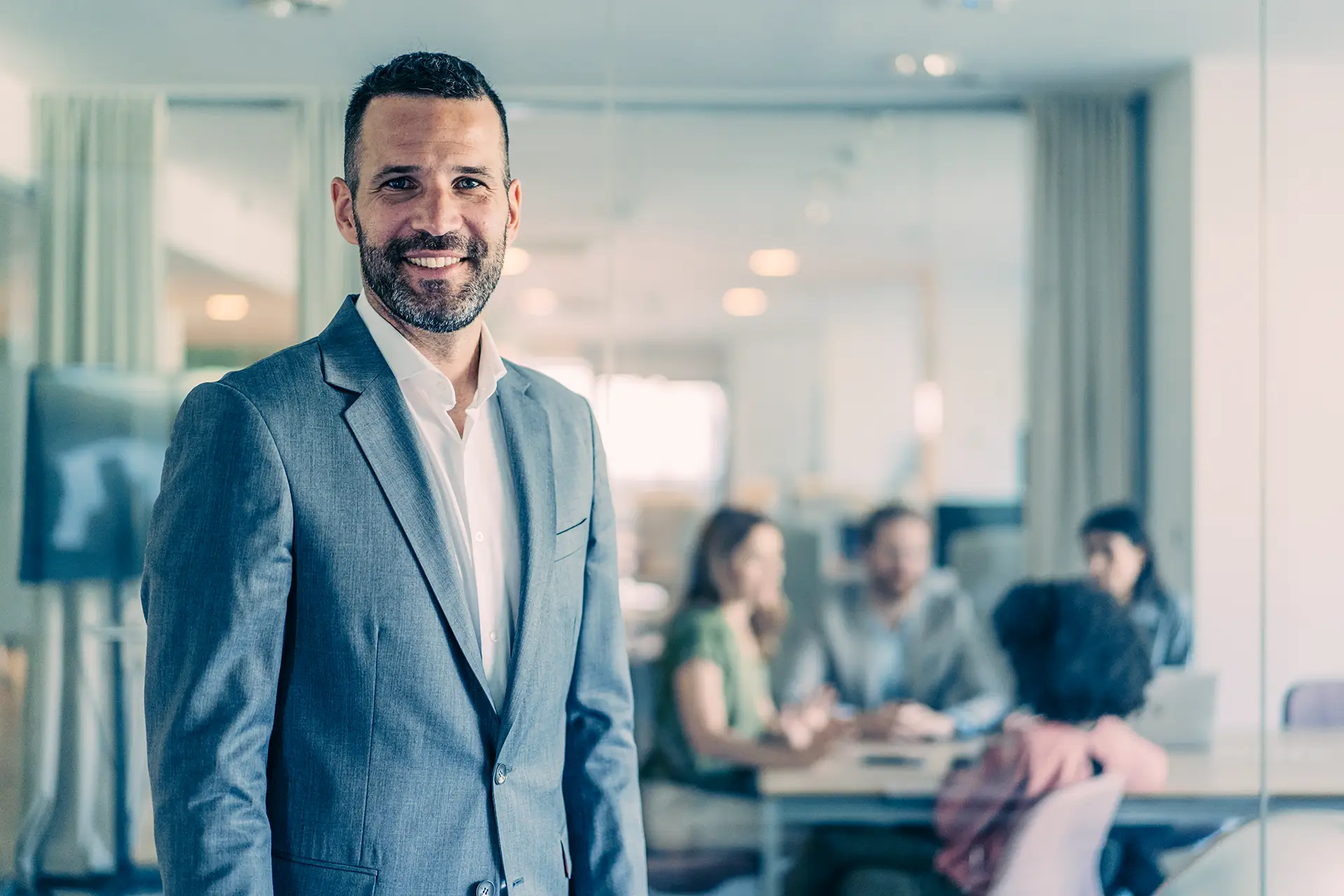 Ein lächelnder Mann in einem grauen Anzug steht in einem modernen Büro. Im Hintergrund sind unscharf Menschen in einem Konferenzraum zu sehen, die an einem Tisch sitzen und diskutieren.