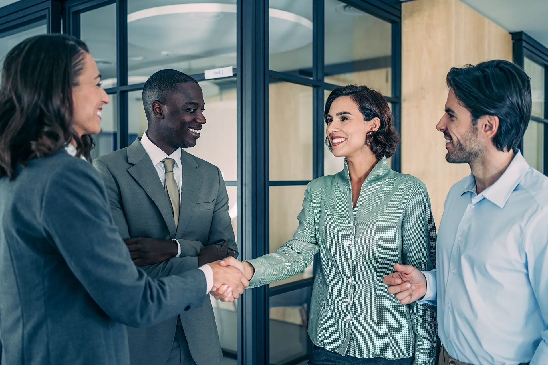 Eine Gruppe von vier Personen in Business-Kleidung steht in einem modernen Büro. Zwei Personen schütteln sich die Hand, während die anderen beiden lächelnd zusehen.